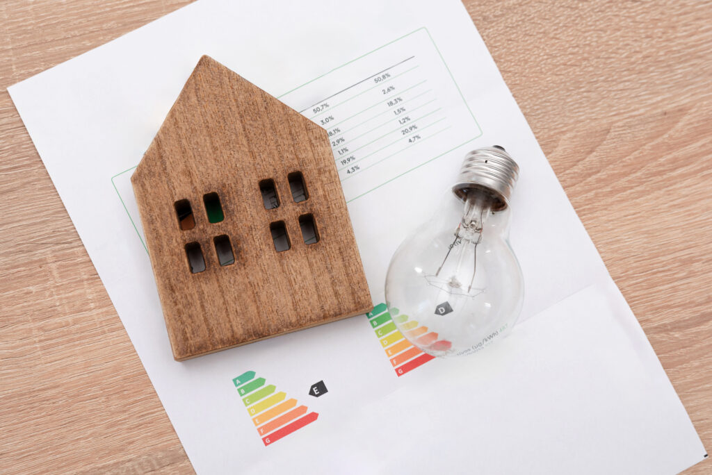 Wooden House Model And And Light Bulb On Energy Efficiency Rating Chart, Electricity Prices And Tax Payments Concept